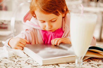 Cute little child girl looks at the book of photography history and drink milk cocktail.