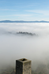 Fototapeta na wymiar A view from an high tower in the little town of San Gimignano, tuscany italy. A sea made of fog until the horizon with light blue mountains in the background and a little town emerging from the mist 