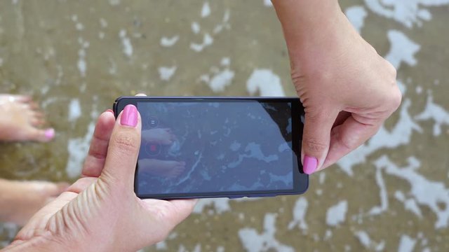 Beach summer vacations concept. Woman relaxing on sandy sea beach using smartphone for taking video selfie for sharing in social media resources. Closeup top view of female hands and legs.