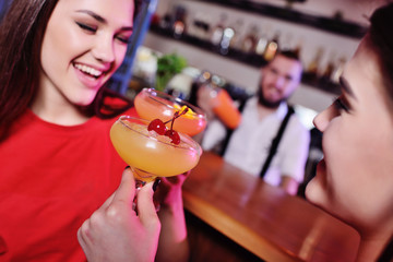 two cute young girlfriend girls are drinking cocktails in a nightclub or bar, having fun and talking
