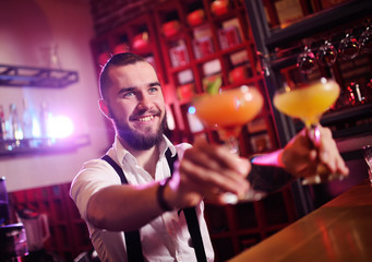 the bartender holding two glasses with an orange alcoholic cocktail smiles against the night club or bar. Bartender Show.