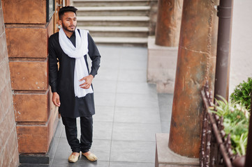 Indian stylish man in black traditional clothes with white scarf posed outdoor.