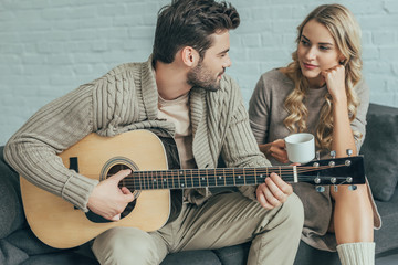 attractive young man playing guitar for girlfriend at home while she sitting on couch with coffee