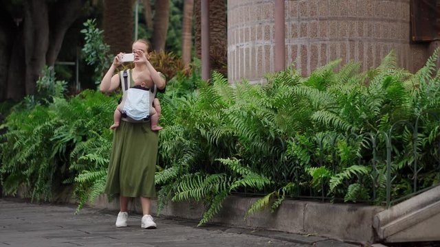 A woman in a dress with a baby in a sling walks along palm trees and shoots video on a mobile phone.