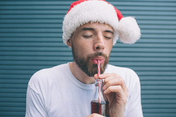 Portrait of bearded man standing and drinking coke through plastic straw. He is keeping eyes closed and enjoying the moment. Guy wears Christmas hat. Isolated on stiped and blue background.