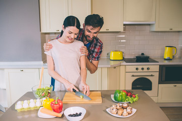 Excited and bearded guy stands behind girl and looks at food. He holds woman on shoulders. Girl looks down as well. She cuts some cheese on desk. Girl is cooking.