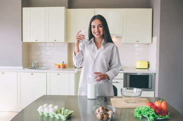 Attractive young woman stands in kitchen and posing. She looks at camera and smiling. Girl holds cup of milk in one hand and big jar of it on the other one.