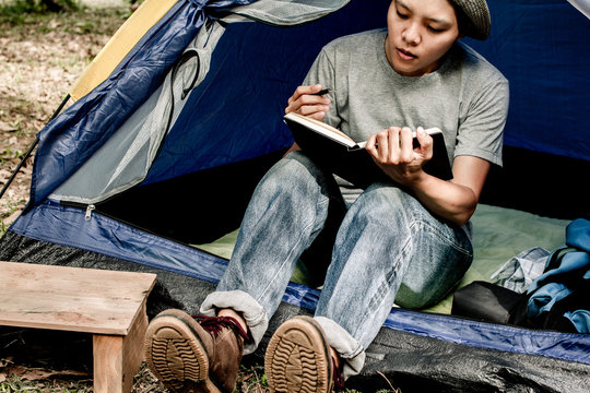 Asian young man sitting is holding a pen writing note of  letter memorize memories on book  in outside the tent. Loneliness camping in forest