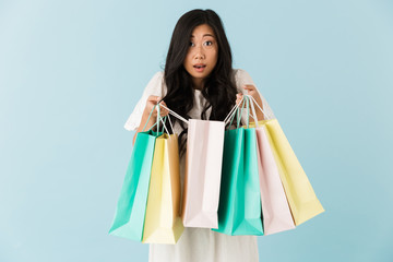 Asian young shocked emotional woman isolated over blue background holding shopping bags.