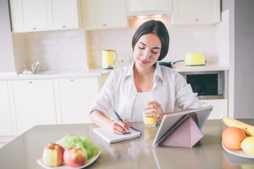 Calm and peaceful young woman sits at table and writes in notebook. She holds glass of juice in hand. There is a tablet standing on table.