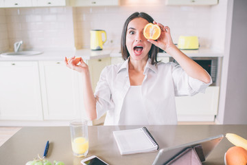 Excited girl is posing on camera. She looks straight. Girl holds piece of orange and hide one eye. There are notebook, tablet, phone, glass of juice and fruit on table.