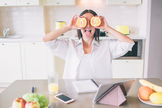 Nice and funny picture of girl hiding her eyes with pieces of orange. She is excited. There are tablet. phone, notebook, glass of juice and plates with fruit at table.