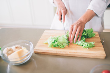 Cut view of girl's hands cutting lettuce on wood board with knife. There is a bowl with boiled chicken and cheese besides it.