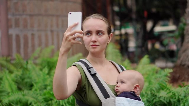 Mom with a baby in a sling holding a smartphone in her hands and communicating via video connection with friends