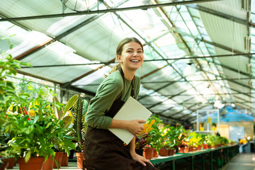 Image of joyful woman gardener smiling on camera, while standing near plants in conservatory with register in hands