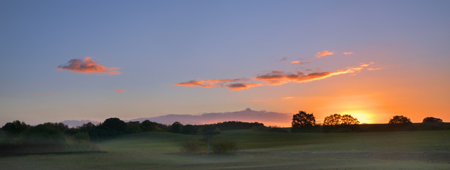 sunrise with glowing clouds over a wide rural landscape with meadows and trees in the morning mist, panorama format with copy space
