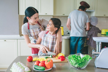 Postive girl are looking at each other and smiling. Mother helps her daughter to cut vegetables in a right way. Dad cooks food with son at stove.