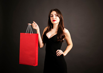 Fototapeta na wymiar Black friday sale concept. Attractive young woman with long brunette hair, smiling, wearing sexy dress, holding red blank shopping bag over black background. Copy space, close up.