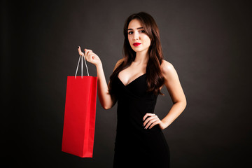Fototapeta na wymiar Black friday sale concept. Attractive young woman with long brunette hair, smiling, wearing sexy dress, holding red blank shopping bag over black background. Copy space, close up.