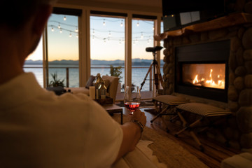 Man tasting red wine inside beautiful home with fireplace and sea view at sunset.