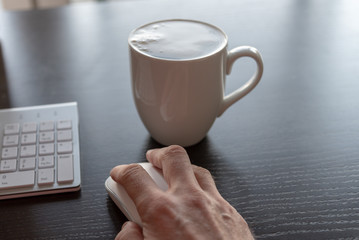 Coffee, keyboard and mouse on desk with hand clicking while working.