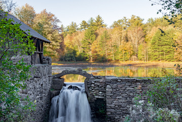 Stone wall and building with waterfall and Autumn trees reflected on Lake in upstate New York.