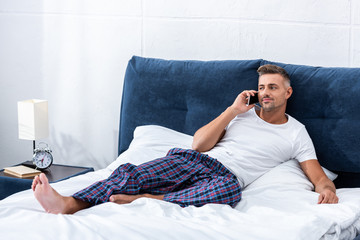 Fototapeta premium cheerful adult man talking on smartphone while laying in bed at home