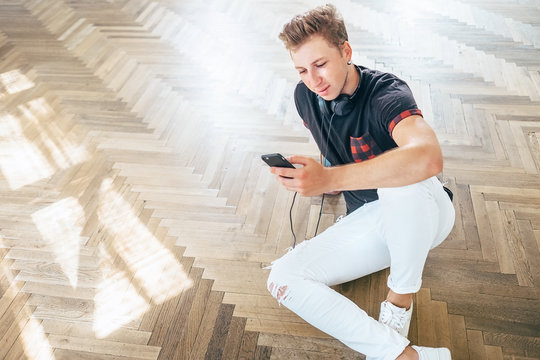 Young man using smartphone with headphones, listen the music and sit on the floor
