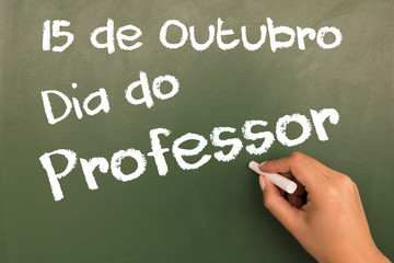 Female hand writing with chalk on green chalkboard October 15 - Teacher's Day (in portuguese).