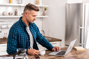 handsome man in checkered shirt holding cup of coffee and using laptop at home