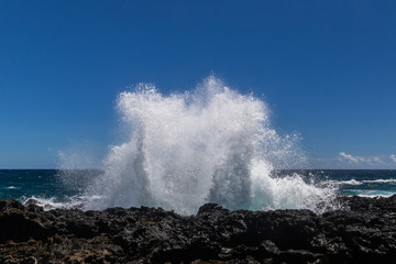 Wave breaking on the rocky coastline of Hawaii's Big Island near South Point. White sea spray thrown into the air; Deep blue Pacific ocean, and blue sky with clouds in the background. 