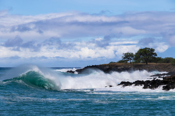 Curling wave cresting and trailing sea spray as it breaks near the shoreline of the Kona coast on Hawaii's Big Island. Behind it is a rocky strip of land with trees. Storm clouds are overhead.