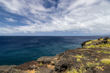 Fototapeta na wymiar View from South Point on the Big Island of Hawaii. This is the southernmost point in the United States. Past the lava rock cliff is the deep blue pacific Ocean; blue sky and clouds in the distance. 