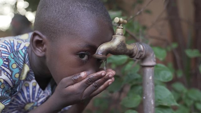 Young African baby boy drinks water from a water tap just outside his rural village. 4K RAW clip, please modify and edit (color grade, stabilize, etc.) in postproduction.