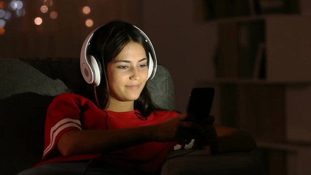 Happy teen selecting song and singing listening to music with headphones and smart phone sitting on a couch