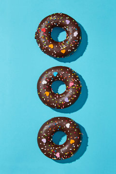 Three chocolate donuts alligned on a blue background viewed from above. Sweet donuts sprinkled with colorful decoration. Top view.  Sharp shadows