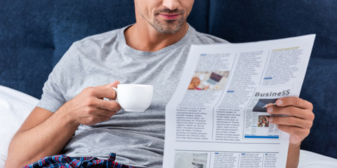Naklejka premium partial view of businessman with cup of coffee reading business newspaper in bed at home
