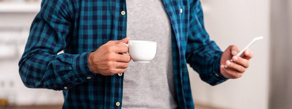 mid section of man in checkered shirt holding cup of coffee and using smartphone at home