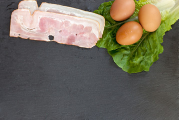 Raw eggs on lettuce and raw bacon on black backround with empty space