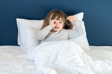 Cute little girl stretching in white bed, yawning and rubbing her eyes, awaking early in the morning