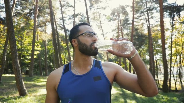 Well-built, bearded man drinking water as having short break during morning run, outdoor shot among tall green trees on fall shiny day