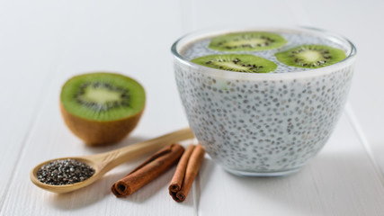 Bowl of Chia seed pudding with kiwi fruit, cinnamon sticks and wooden spoon on white table.