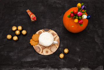 Coffee with white creamy foam on a black table. Nearby are a souvenir red wooden bird, candy wrapped in a golden wrapper, cookies, and flowers in an orange vase