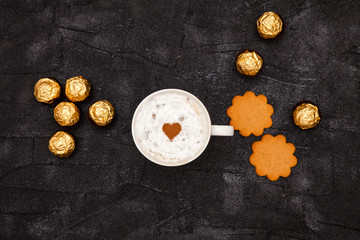 Coffee with white creamy foam and heart-shaped chocolate on a black table. Next to the round candy in a golden wrapper and cookies
