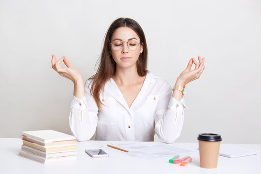People and spirituality concept. Relaxed brunette young woman poses at workplace in mudra sign, enjoys peaceful atmosphere, pull herself together and prepares for work, drinks takeaway coffee