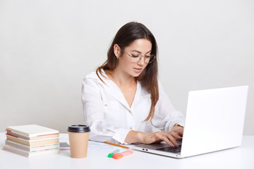 Busy freelancer recieves email, types feedback on laptop computer, wears round glasses, dressed in white shirt, surrounded with books, isolated over white background. People and career concept