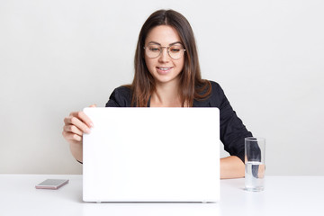 Satisfied young female sits in front of opened laptop computer, watches webinar, thinks about creating new web design, wears round spectacles for good vision, drinks water, sits indoor alone.