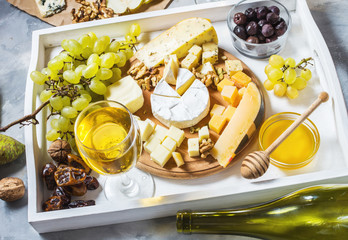Different types of cheese on wooden board, olive, fruits, almond and wine glasses on white tray