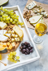 Different types of cheese on wooden board, olive, fruits, almond and wine glasses on white tray