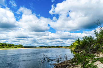 A wide river flows along a diagonal bank with a forest under an incredibly beautiful blue sky with large clouds that are reflected in the water. Belarusian Polesie. Pripyat National Park